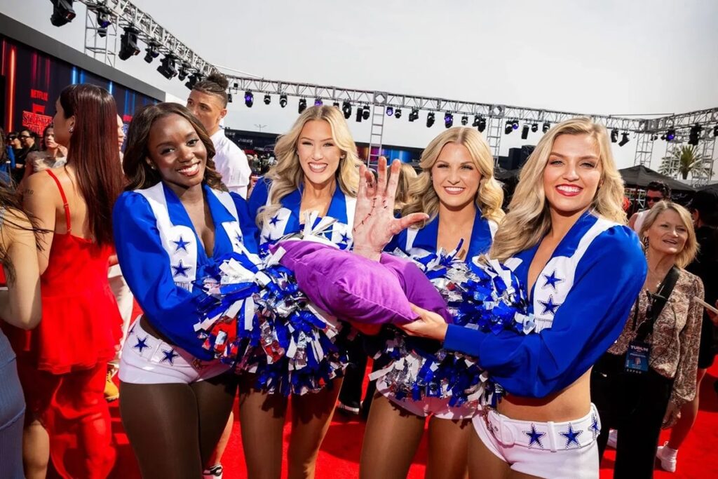 Dallas Cowboys Cheerleaders smiling on the red carpet, holding a prop hand from Stranger Things